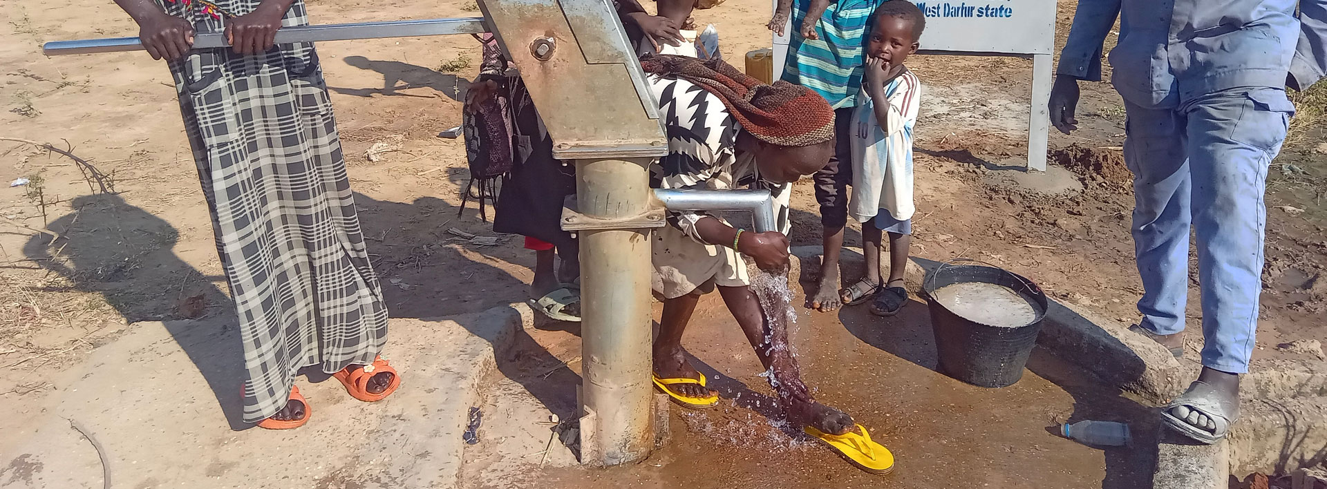 Photo of several sudanese people standing around a water pump with a child bending down and drinking from the pump