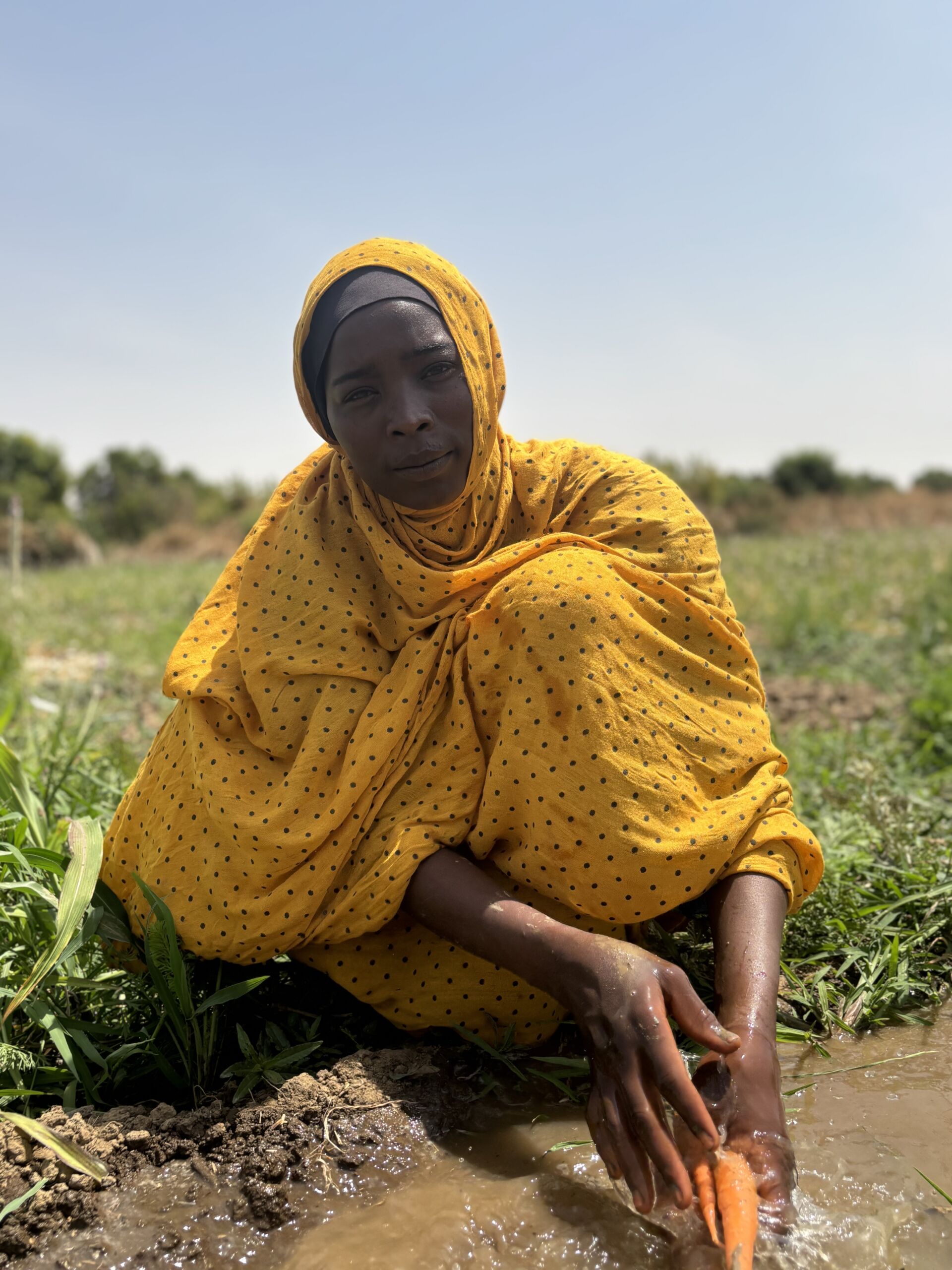 Mission East og vores lokale partner har uddelt frø, så de flygtninge, der bor i Dougui-lejren, kan dyrke afgrøder. På billedet ses en ung kvinde, der sidder i marken iført gule klæder. I det lille vandløb renser hun de nyligt plukkede gulerødder fra hendes have i Dougui-lejren.