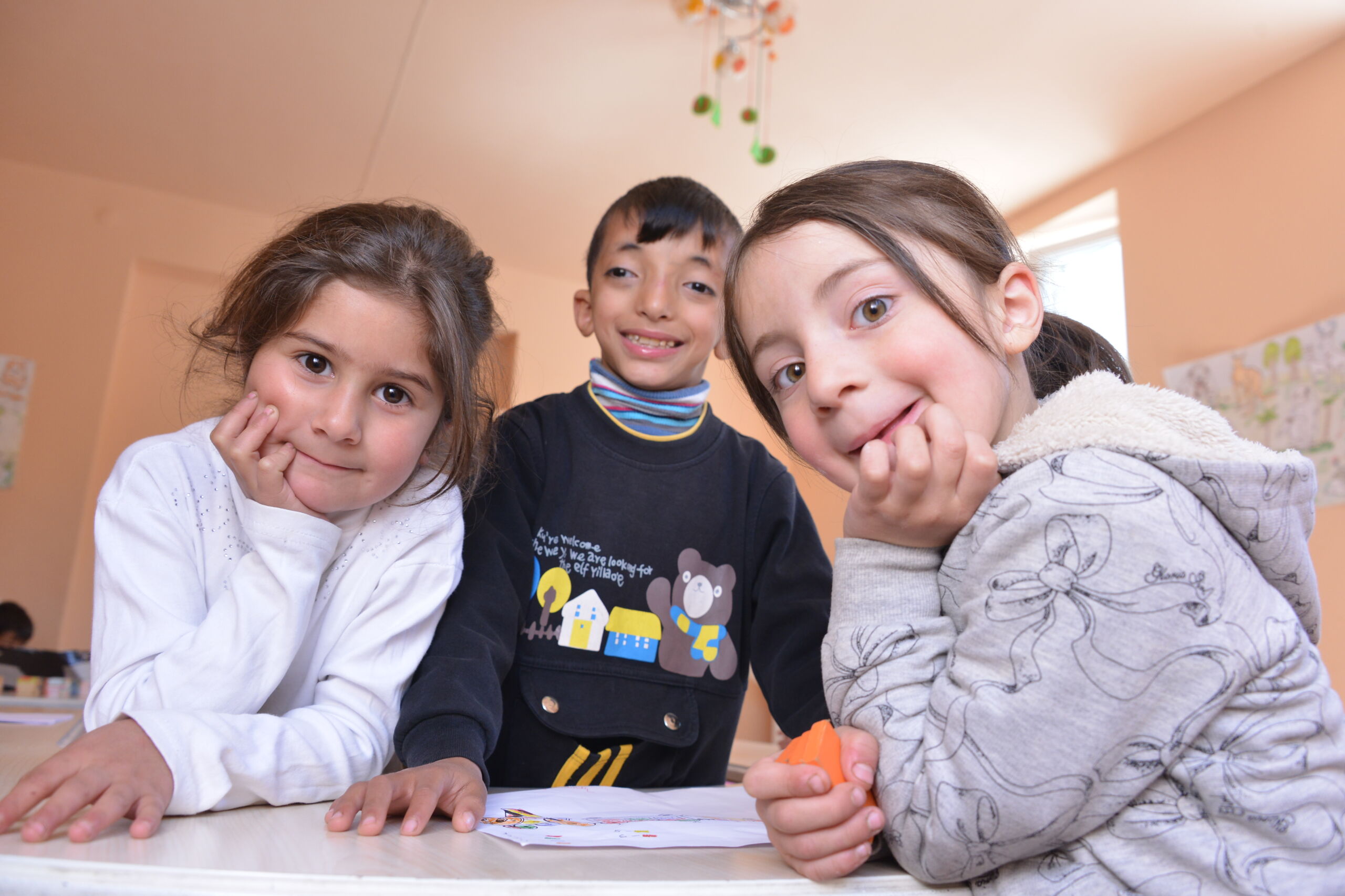 Tre børn smiler nysgerrigt og fulde af liv ind i kameraet – et øjeblik af glæde og nærvær.